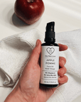 Hand holding a bottle of 'Dry Skin Love Apple 5% Vitamin E Face Oil' over a white cloth on a white counter with a red apple in the background, luxury face oil
