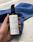 Hand holding bottle of 'Dry Skin Love Blueberry Oil Cleanser for Acne Prone Skin' on blue cloth on a white counter, luxury face cleanser