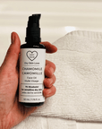 Hand holding a bottle of 'Dry Skin Love Chamomile 1% Bisabolol Face Oil' for Sensitive Skin on a white cloth on a white counter, luxury face oil