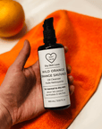 Hand holding a bottle of 'Dry Skin Love' Wild Orange Oil Cleanser on an orange towel with oranges in the background, luxury face cleanser