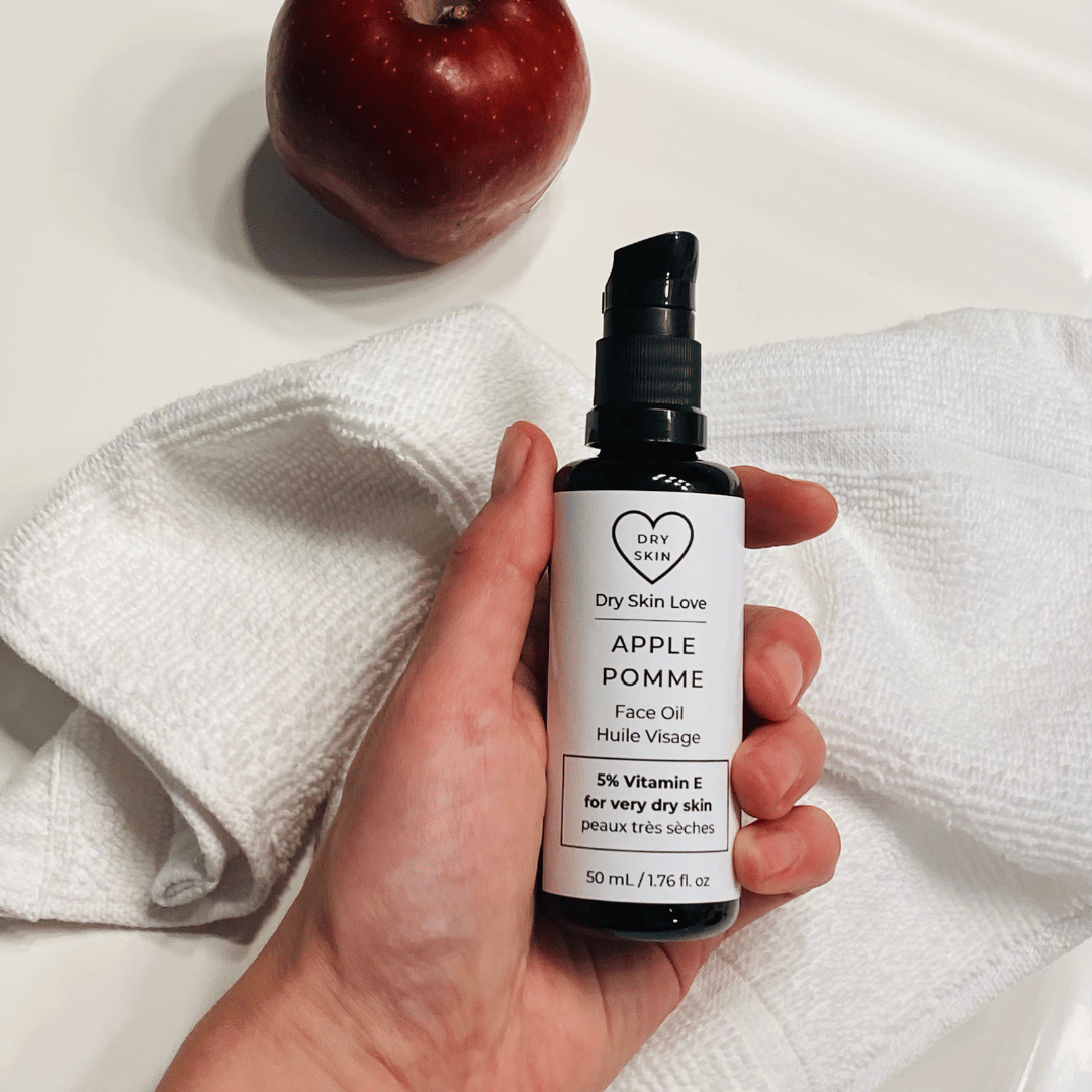 Hand holding a bottle of 'Dry Skin Love Apple 5% Vitamin E Face Oil' over a white cloth on a white counter with a red apple in the background, luxury face oil