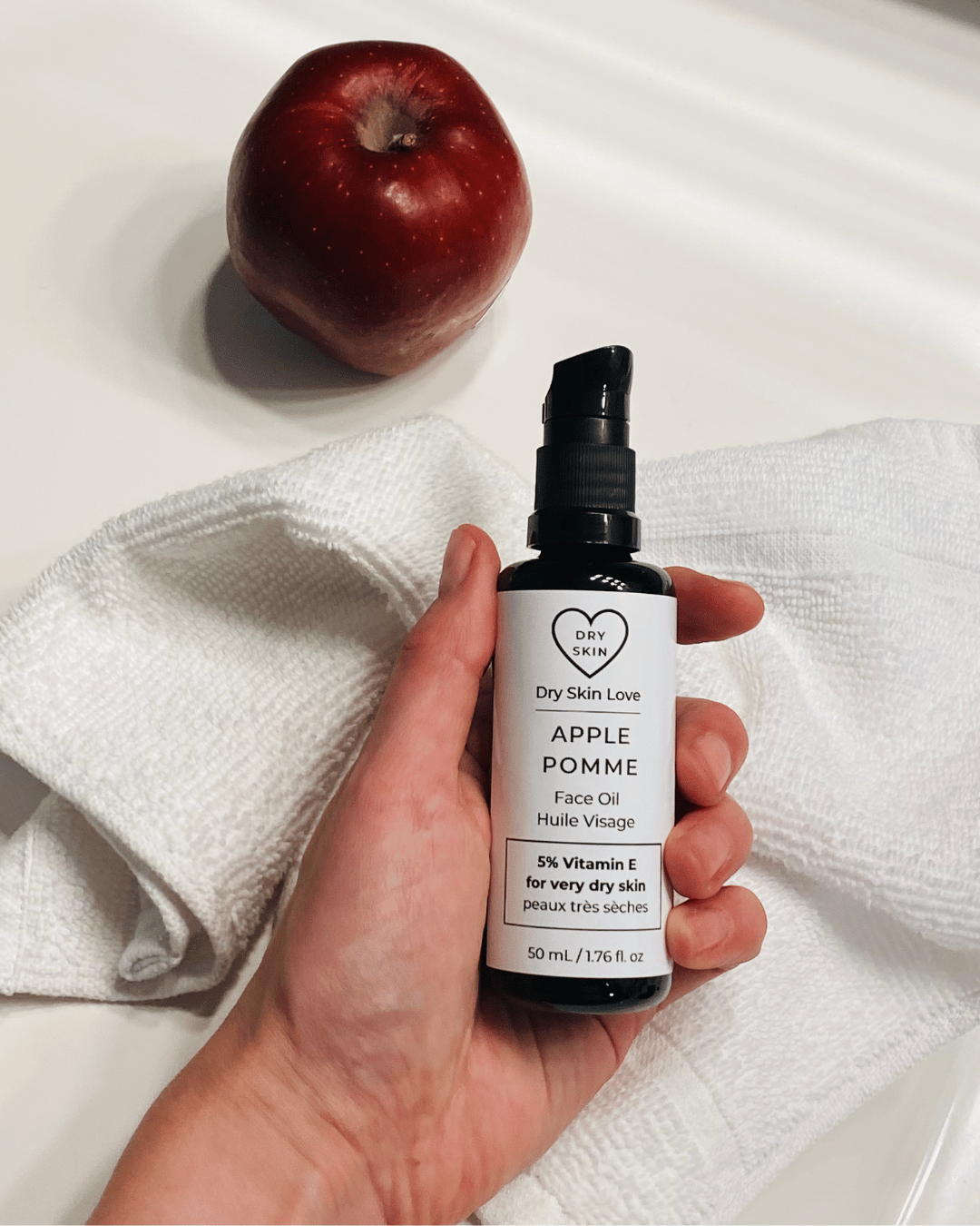 Hand holding a bottle of 'Dry Skin Love Apple 5% Vitamin E Face Oil' over a white cloth on a white counter with a red apple in the background, luxury face oil