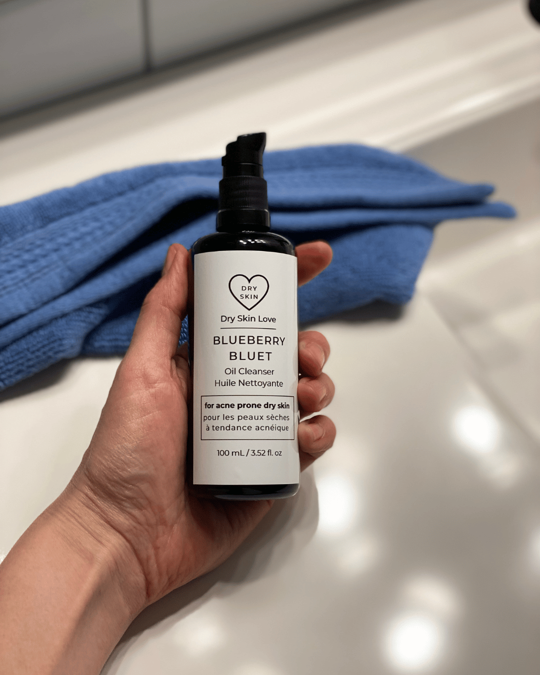 Hand holding a bottle of Blueberry Oil Cleanser with a blue towel in the background, luxury face cleanser