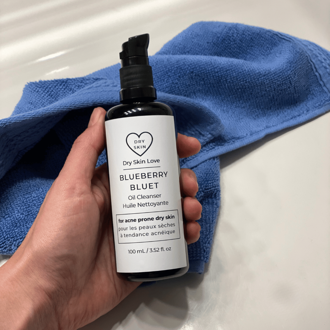 Hand holding bottle of 'Dry Skin Love Blueberry Oil Cleanser for Acne Prone Skin' on blue cloth on a white counter, luxury face cleanser