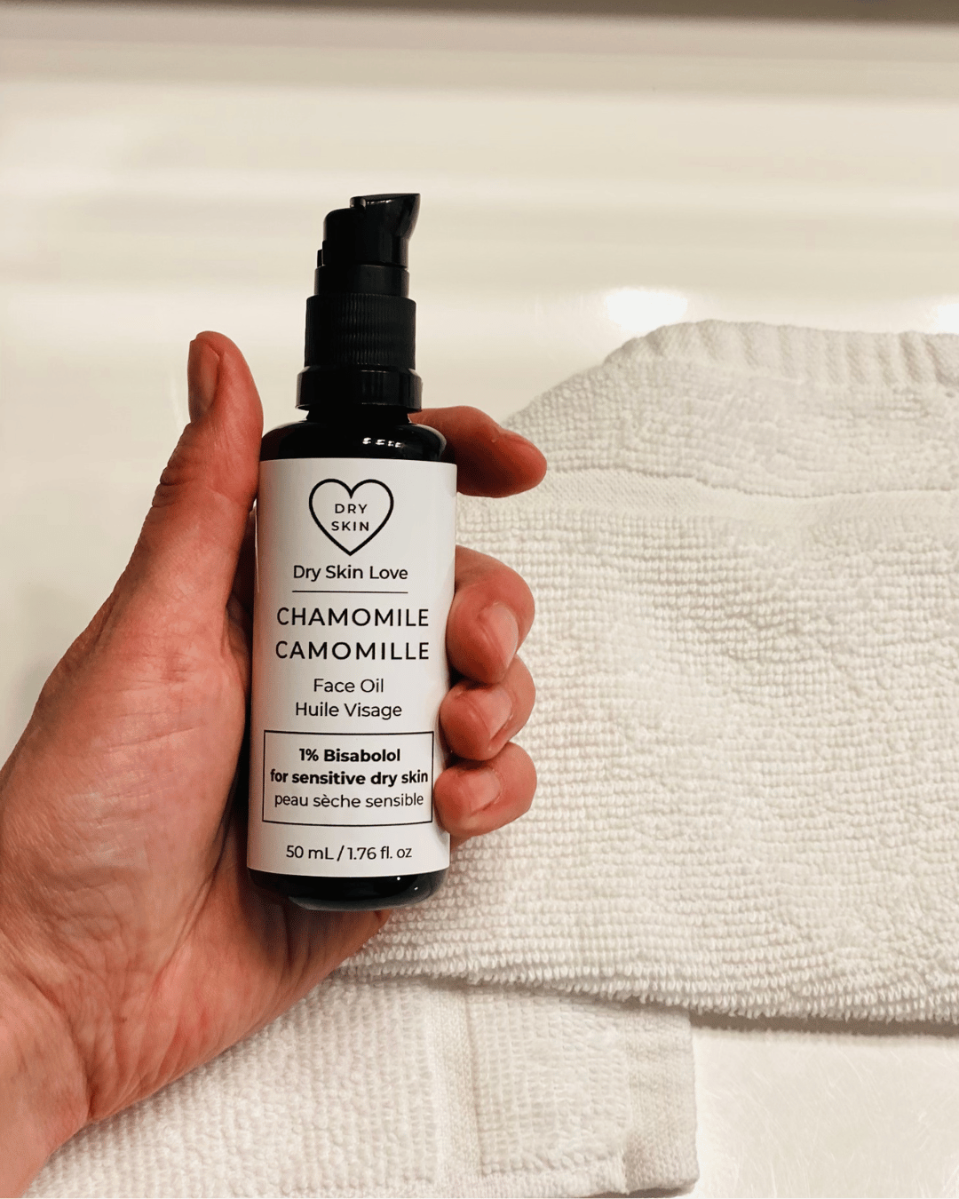Hand holding a bottle of 'Dry Skin Love Chamomile 1% Bisabolol Face Oil' for Sensitive Skin on a white cloth on a white counter, luxury face oil