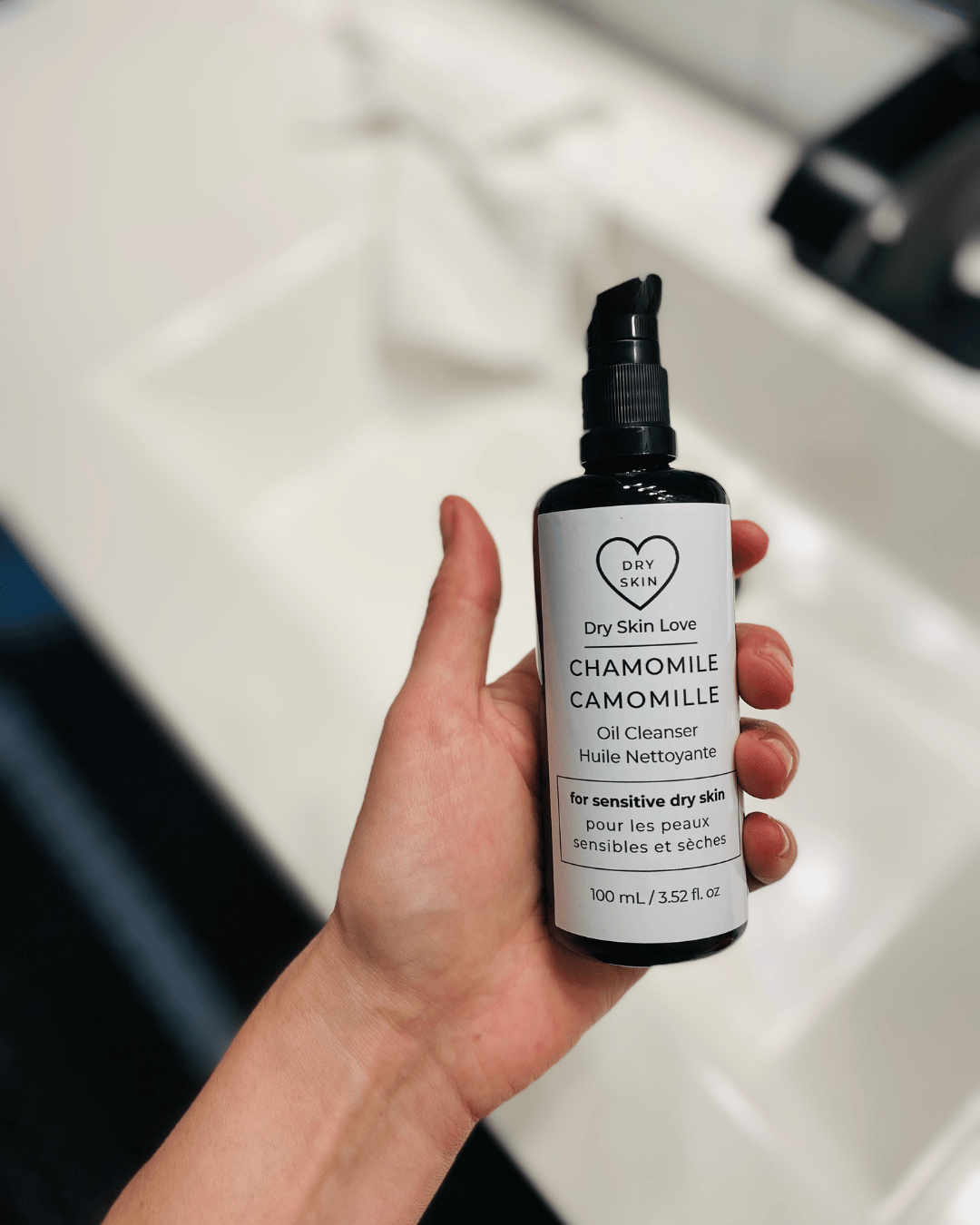 Hand holding a bottle of 'Chamomile Oil Cleanser for Dry Sensitive Skin' with a white sink in the background, luxury face cleanser