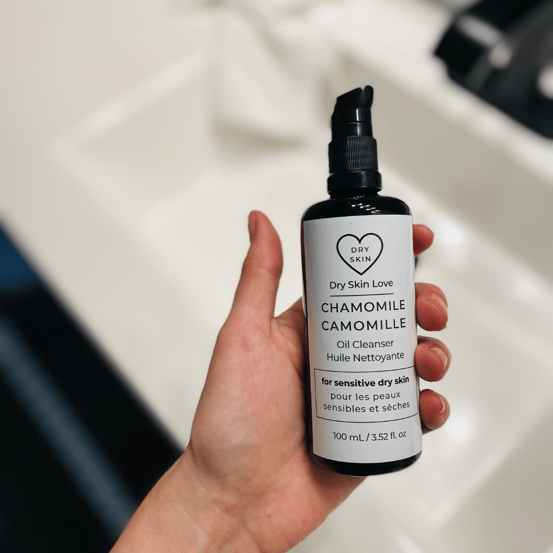 Hand holding a bottle of 'Dry Skin Love Chamomile Oil Cleanser for Sensitive Skin' over a white sink, Luxury Face Cleanser 