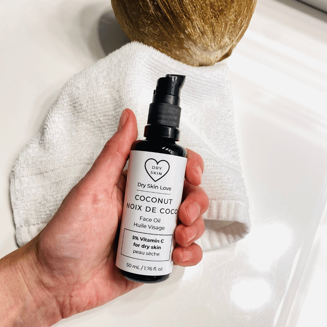 Hand holding a bottle of &#39;Dry Skin Love Coconut 5% Vitamin C Face Oil&#39; over a white cloth on a white counter with a coconut in the background, luxury face oil