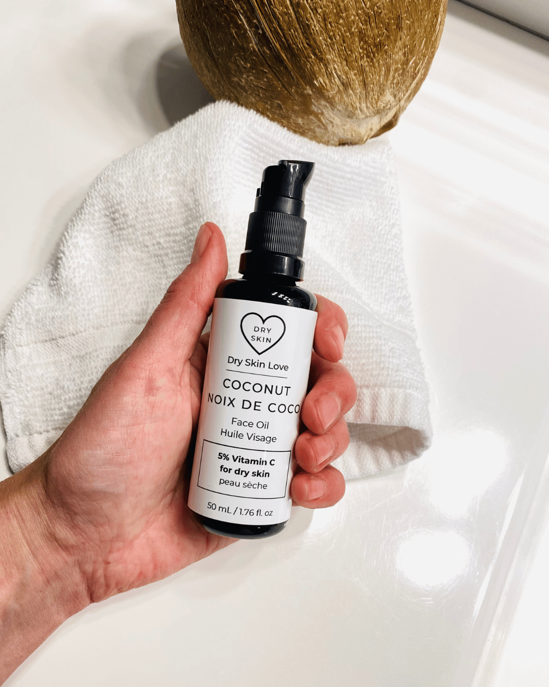 Hand holding a bottle of 'Dry Skin Love Coconut 5% Vitamin C Face Oil' over a white cloth on a white counter with a coconut in the background, luxury face oil