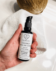 Hand holding a bottle of 'Dry Skin Love Coconut 5% Vitamin C Face Oil' over a white cloth on a white counter with a coconut in the background, luxury face oil