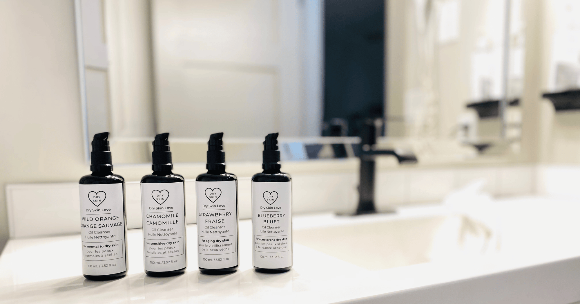 Four Miron violet glass bottles of Dry Skin Love oil cleansers on a bathroom counter with sink and mirror in the background, luxury skincare for dry skin.