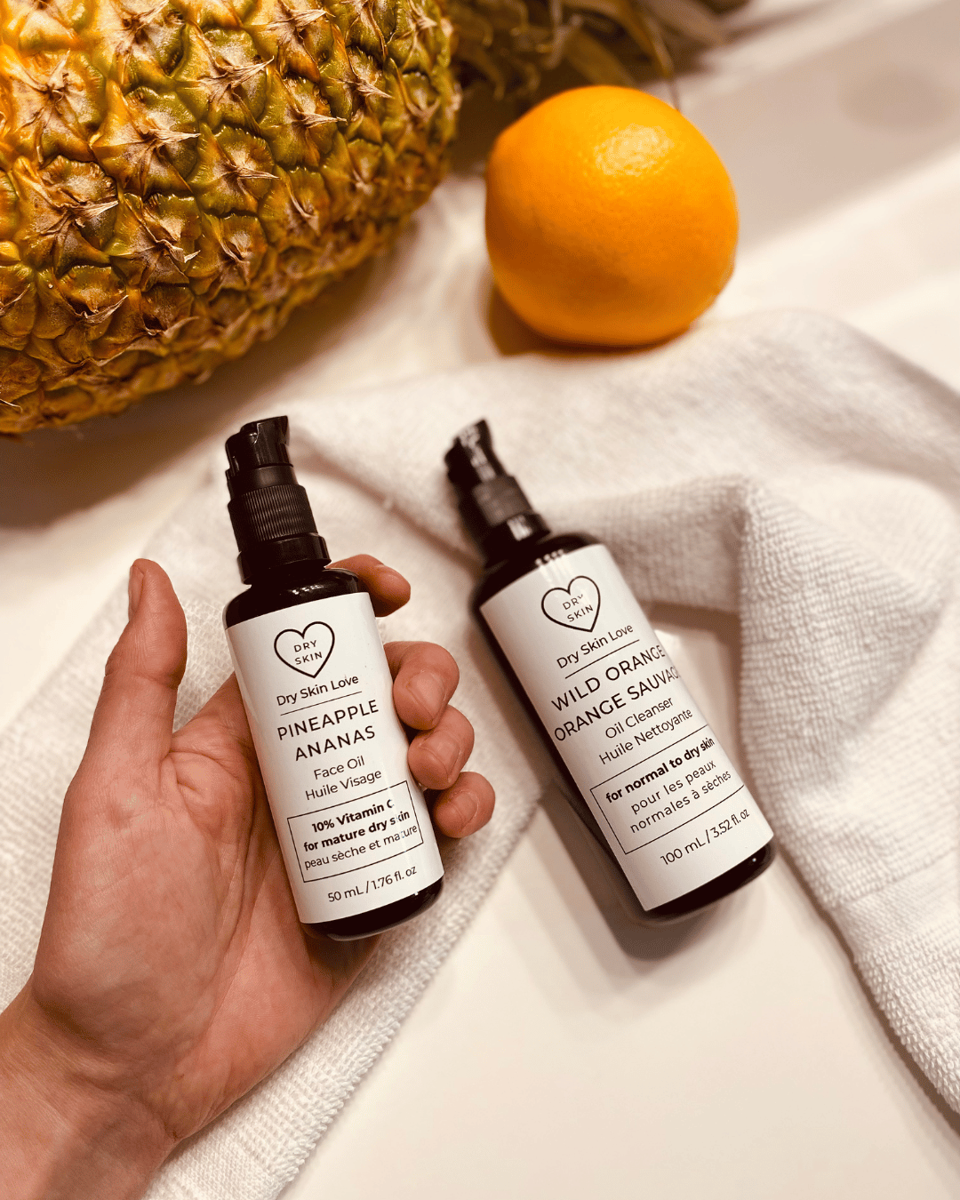Two glass bottles of Dry Skin Love skincare products—Pineapple 10% Vitamin C Face Oil and Wild Orange Oil Cleanser—resting on a white cloth on a white countertop, with a fresh pineapple and orange in the background.