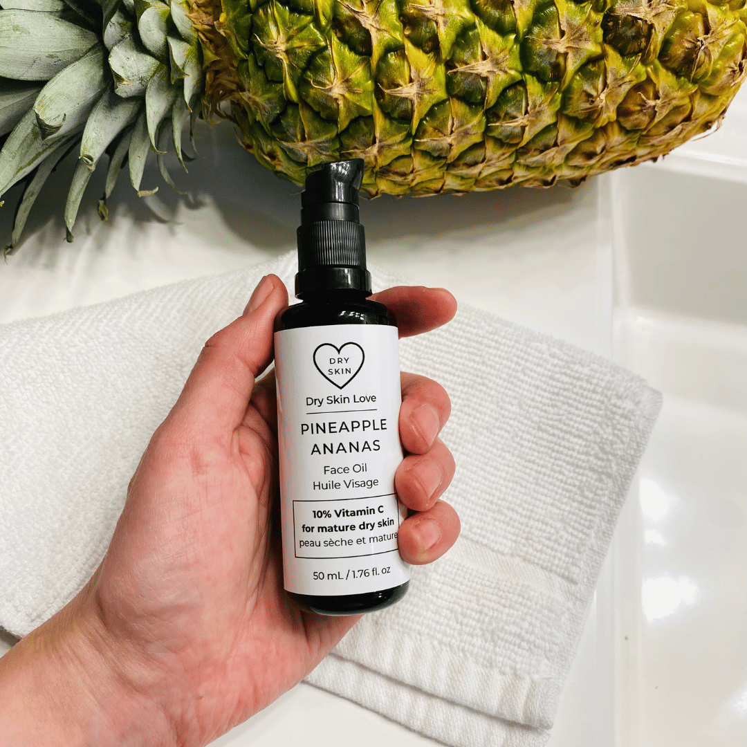 Hand holding a bottle of 'Dry Skin Love Pineapple 10% Vitamin C Face Oil' over a white cloth on a white counter with a pineapple in the background, luxury face oil