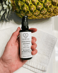Hand holding a bottle of 'Dry Skin Love Pineapple 10% Vitamin C Face Oil' over a white cloth on a white counter with a pineapple in the background, luxury face oil