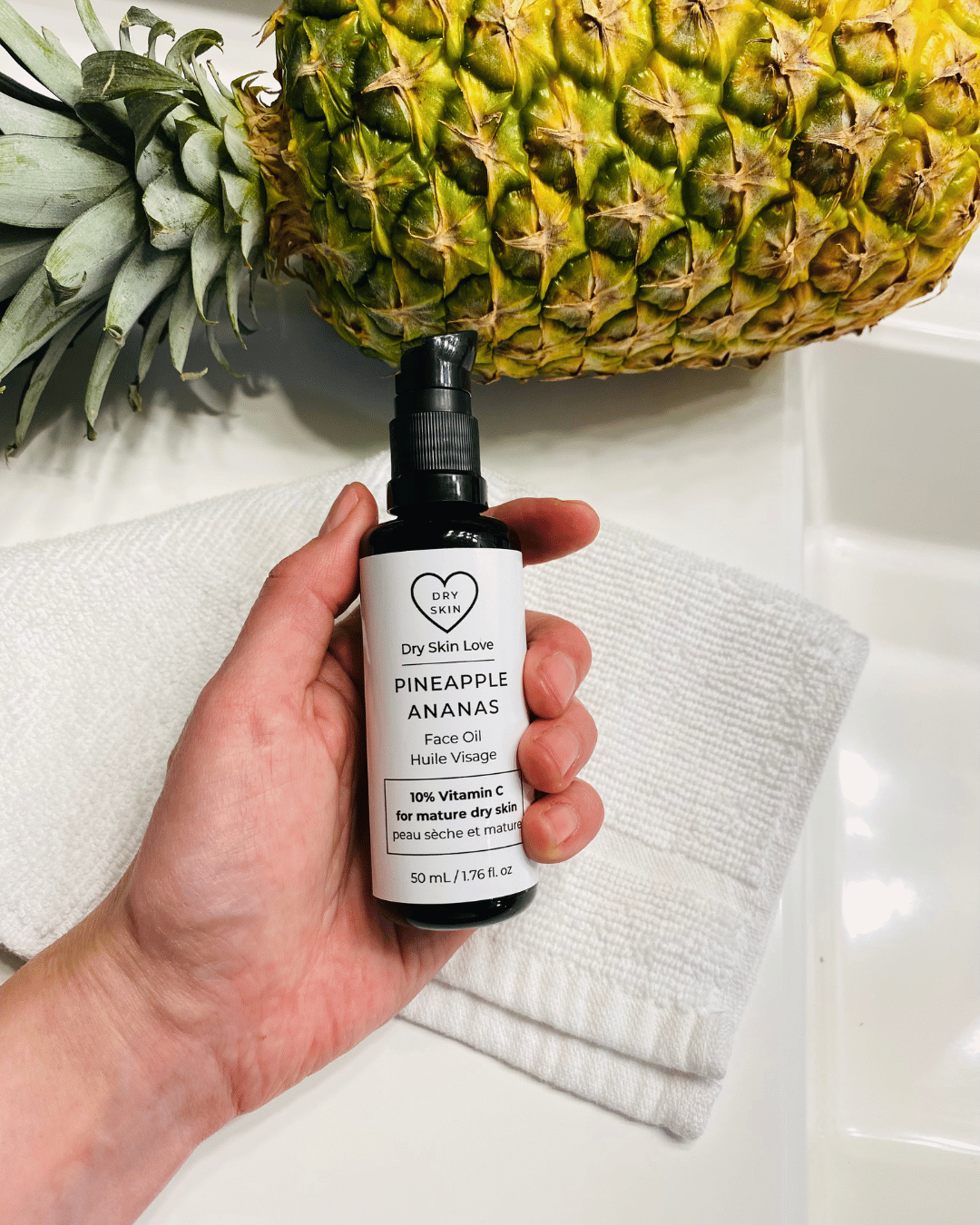 Hand holding a bottle of 'Dry Skin Love Pineapple 10% Vitamin C Face Oil' over a white cloth on a white counter with a pineapple in the background, luxury face oil