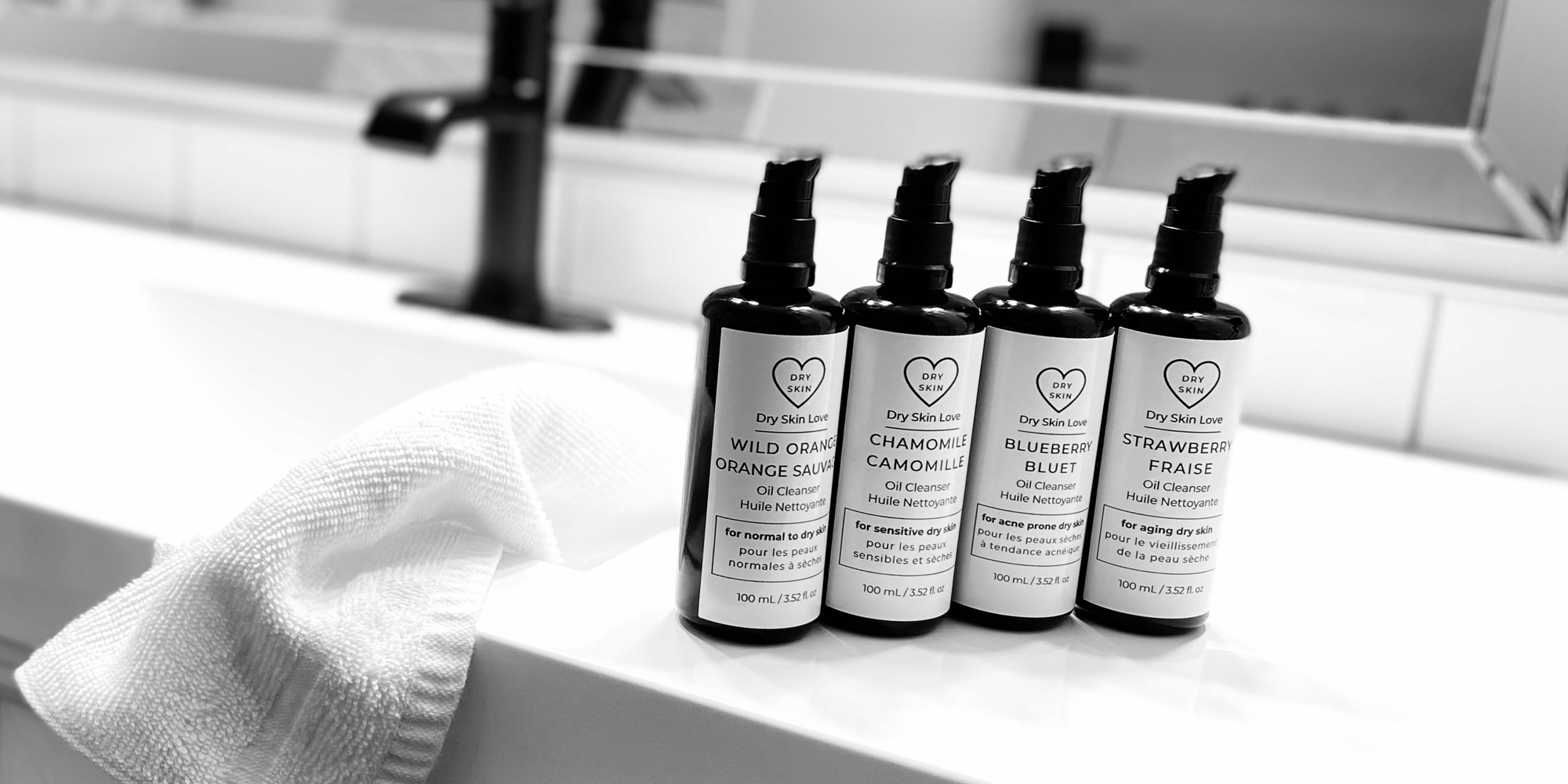 Four Miron violet glass bottles of Dry Skin Love oil cleansers displayed on on a bathroom counter with sink and mirror in the background, luxury skincare for dry skin.