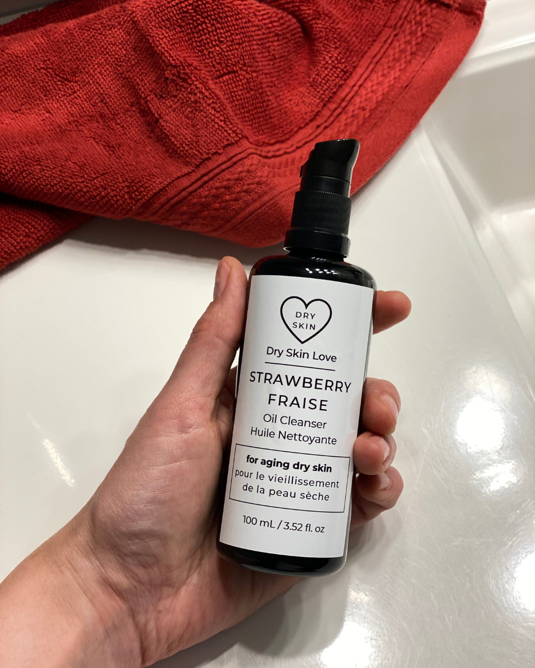 Hand holding a bottle of 'Dry Skin Love Strawberry Oil Cleanser' with a red towel in the background, Luxury Face Cleanser for Dry Aging Skin