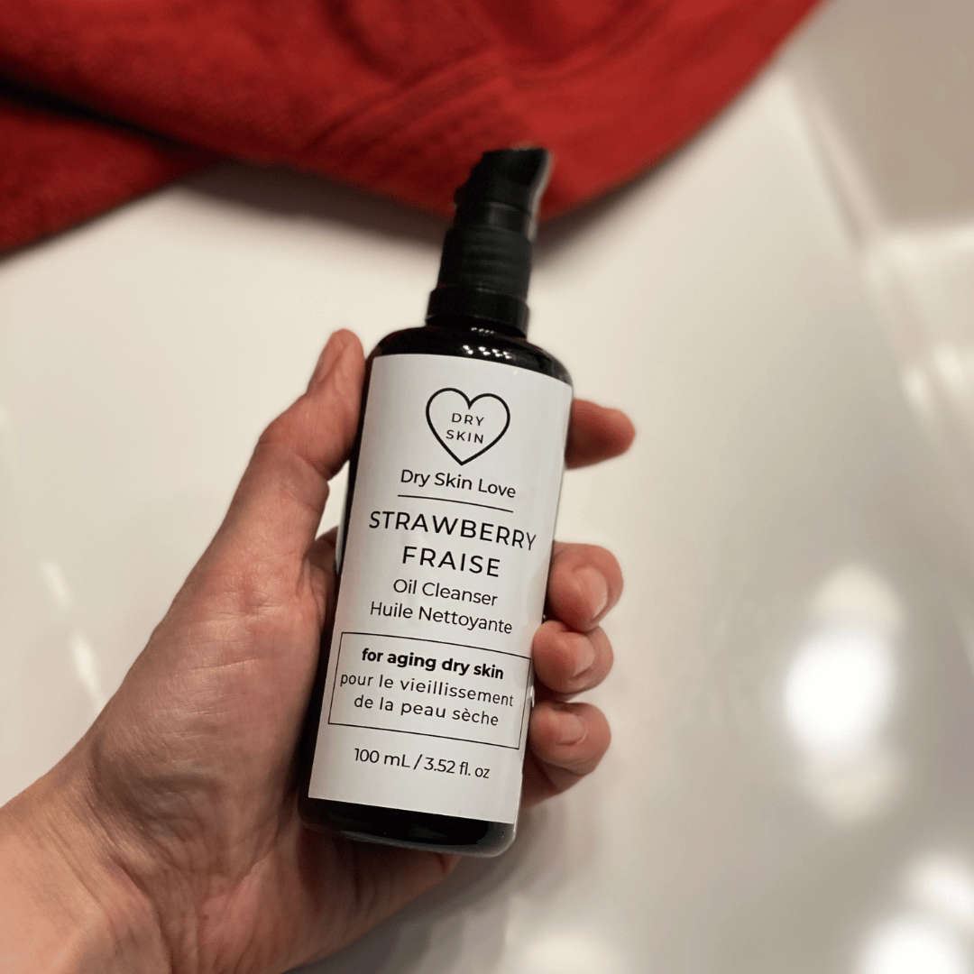 Hand holding bottle of 'Dry Skin Love Strawberry Oil Cleanser for Mature Skin' with red cloth in background on a white counter, luxury face cleanser