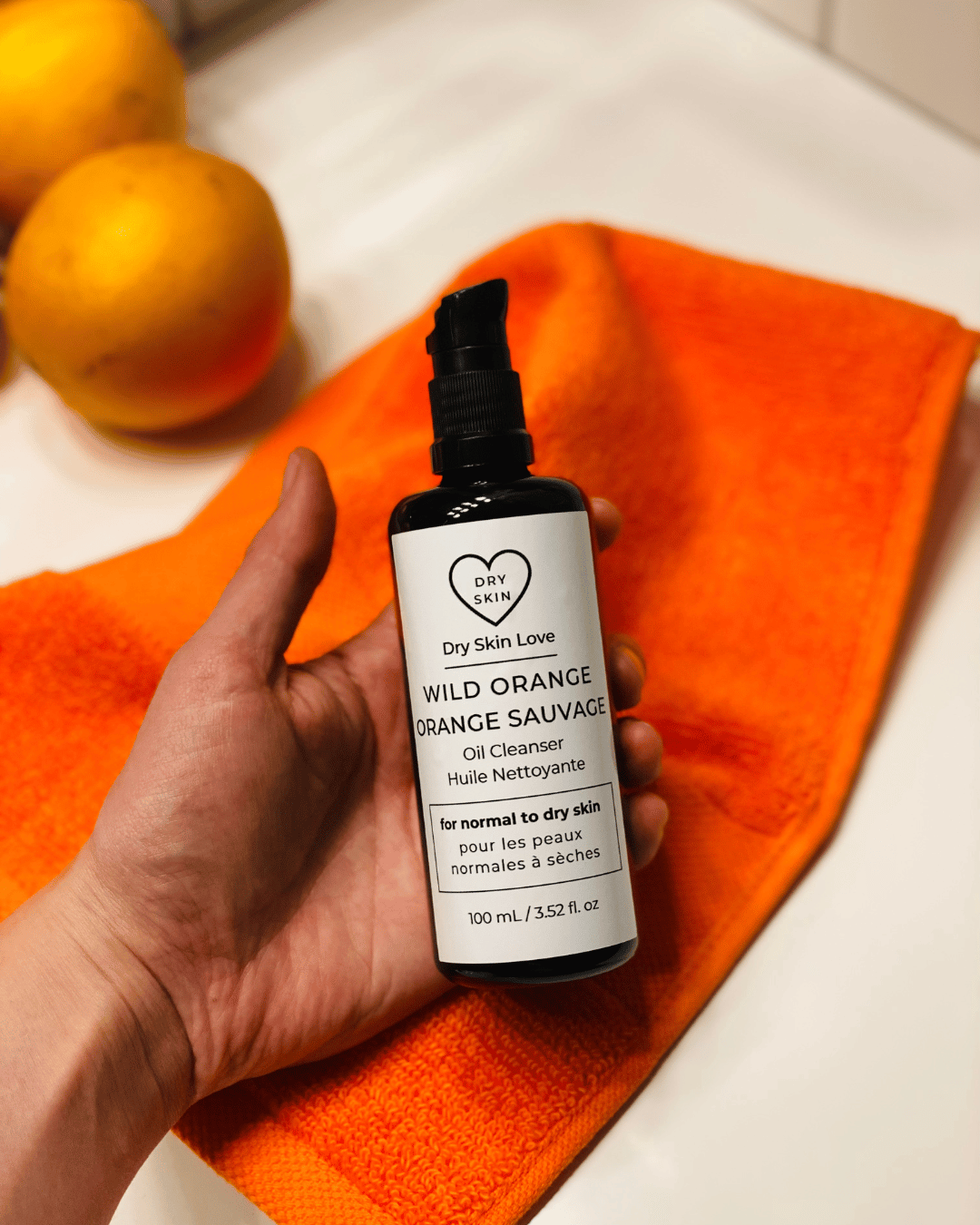 Hand holding a bottle of Dry Skin Love Wild Orange Oil Cleanser on an orange towel with oranges in the background.