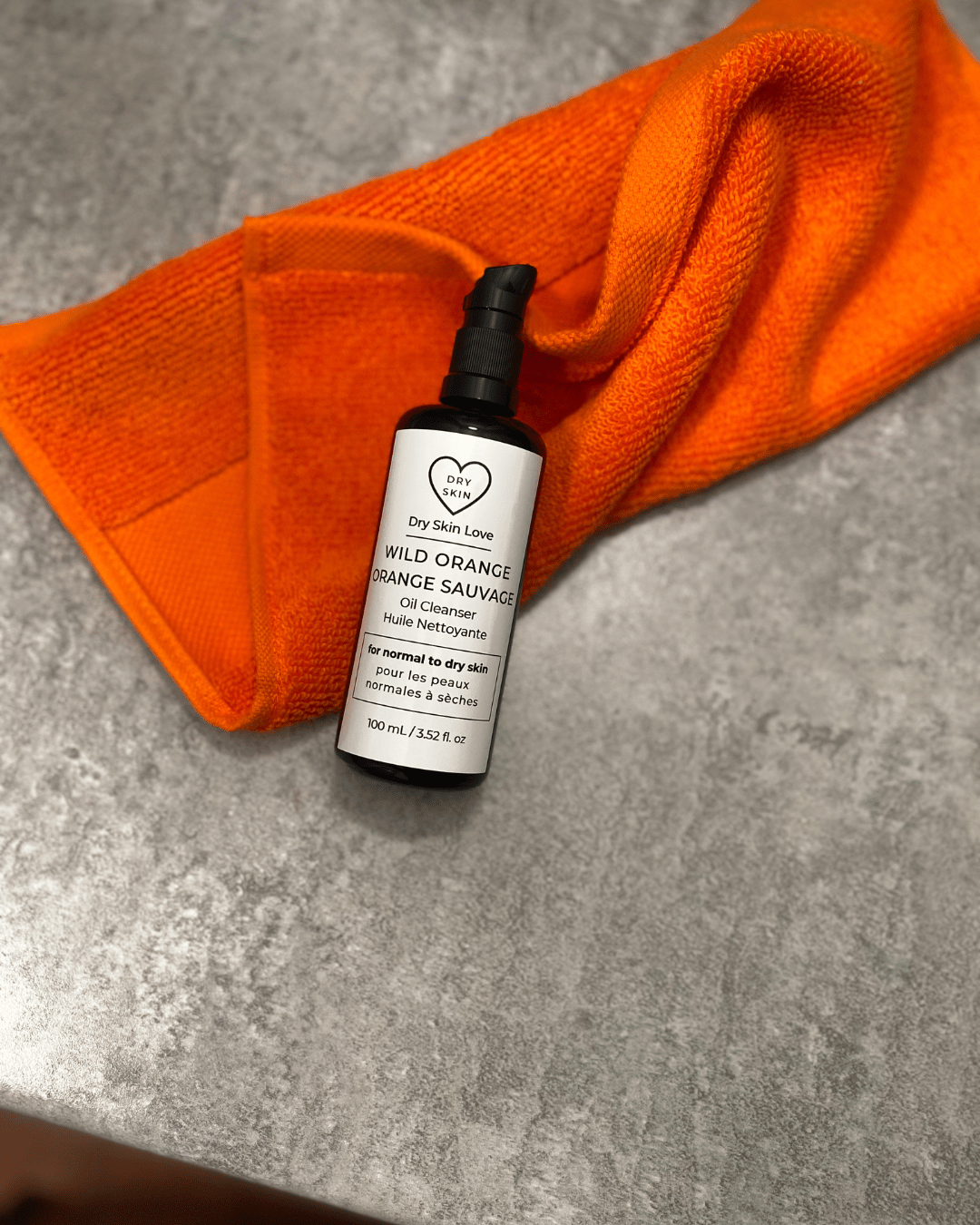 Miron violet glass bottle of Dry Skin Love Wild Orange Oil Cleanser resting on an orange towel, with a fresh orange fruit in the background, luxury skincare for dry skin.