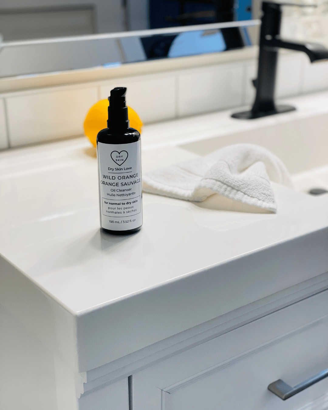 Bottle of Dry Skin Love Wild Orange Oil Cleanser on a bathroom counter with a mirror in the background, luxury face cleanser