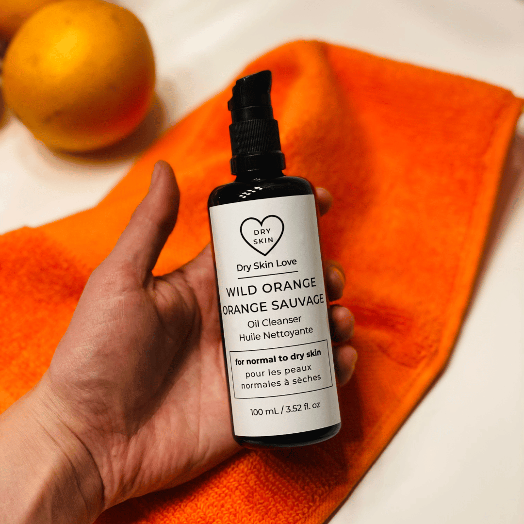 Hand holding a bottle of 'Dry Skin Love' Wild Orange Oil Cleanser on an orange towel with oranges in the background, luxury face cleanser